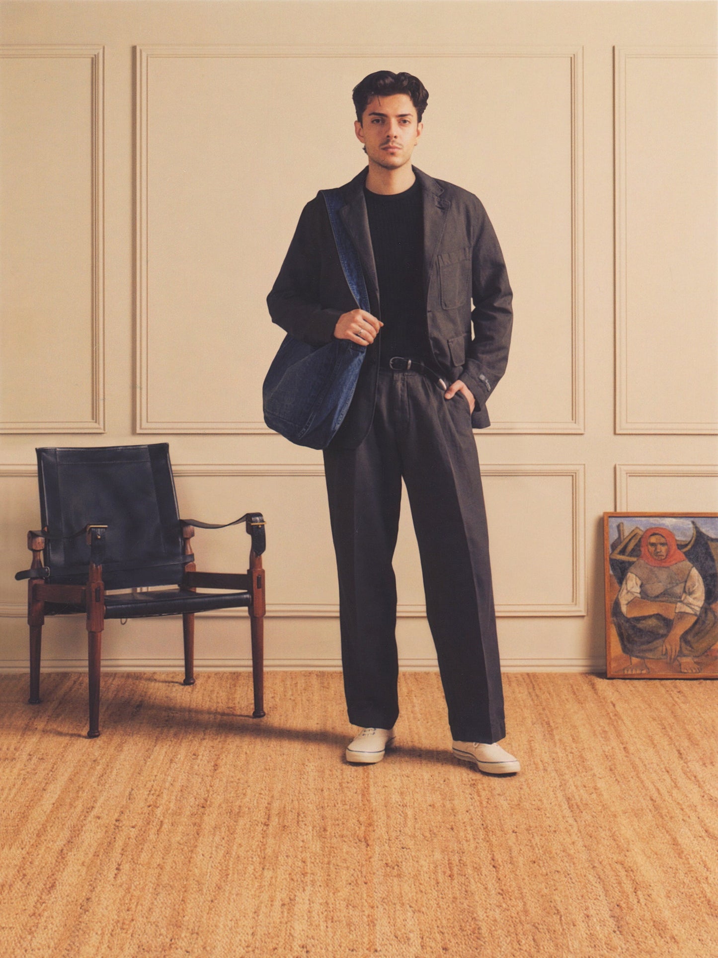 Man in a stylish outfit standing in a room with a chair and painting.